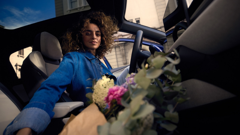 A woman is sitting in the driver's seat of a blue VW ID. Polo and reaches for a bouquet of flowers on the passenger seat.
