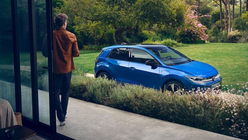 A view through an open terrace door into a green garden, a blue VW ID. Polo stands in the driveway.