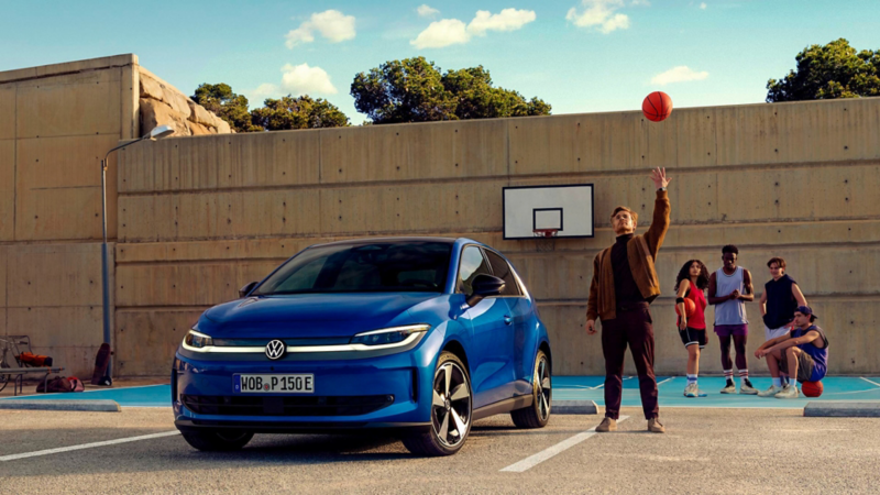 Ein Mann wirft einen Basketball in die Luft, neben ihm parkt ein blauer VW ID. Polo vor einem Basketballplatz.