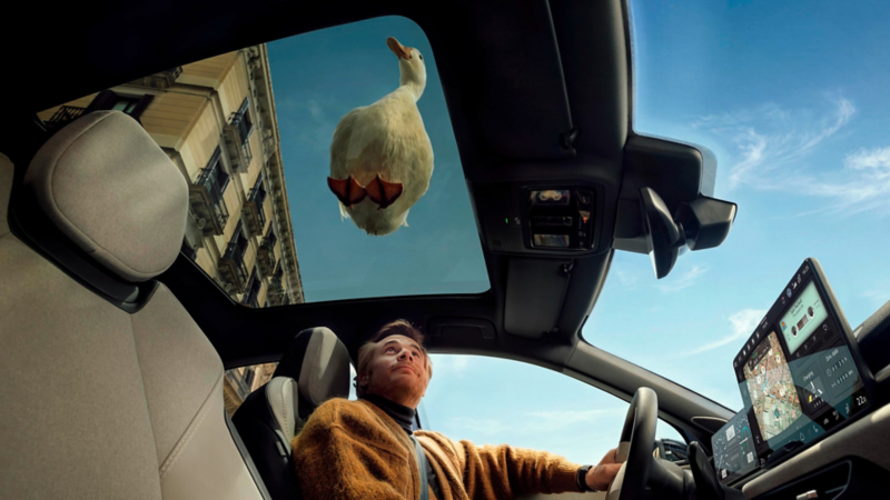 Un homme regarde à travers le toit panoramique de la VW ID. Polo, avec un canard blanc posé sur le toit