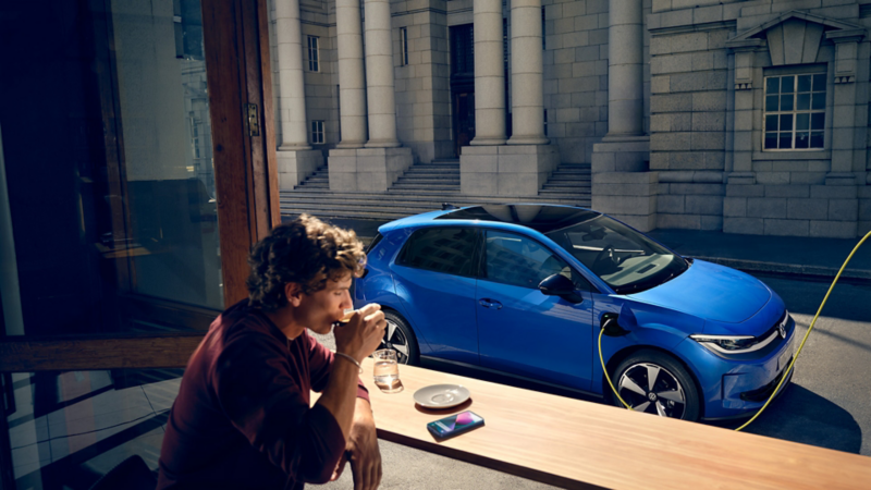 Ein Mann sitzt in einem Café, durch ein offenes Fenster ist der blaue VW ID. Polo Style zu sehen, der an einer Ladestation lädt.