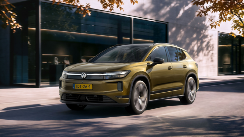 An olive-bronze VW ID. Tiguan drives along a road in front of a modern building.