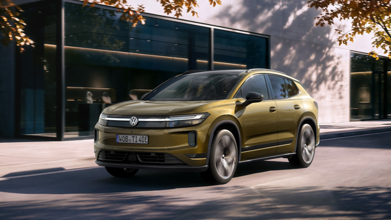 An olive-bronze VW ID. Tiguan drives along a road in front of a modern building.
