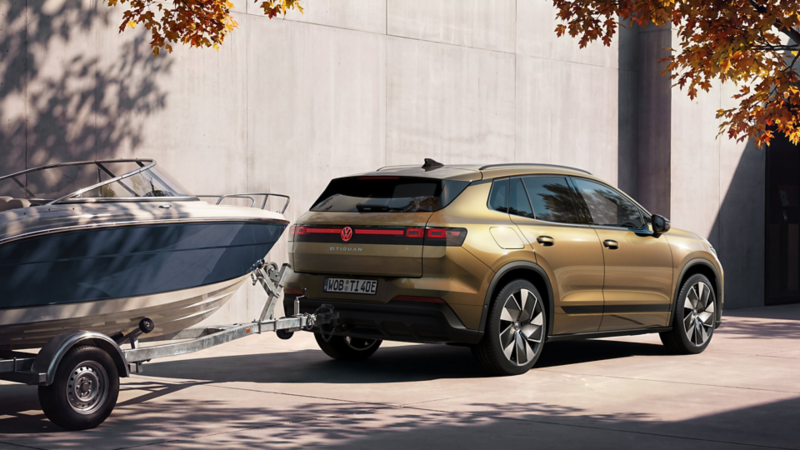 An olive-bronze VW ID. Tiguan is parked in front of a modern building, towing a trailer carrying a boat.