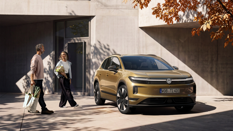 Un homme et une femme chargés de leurs achats s'approchent d'un VW ID. Tiguan de couleur bronze olive, garé devant un bâtiment moderne.