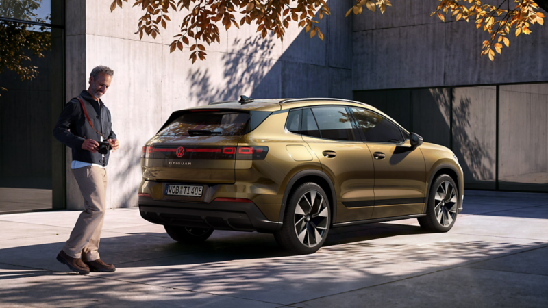 Rear side view of an olive-bronze VW ID. Tiguan Style parked in front of a modern building. A man holding a camera walks toward the vehicle.