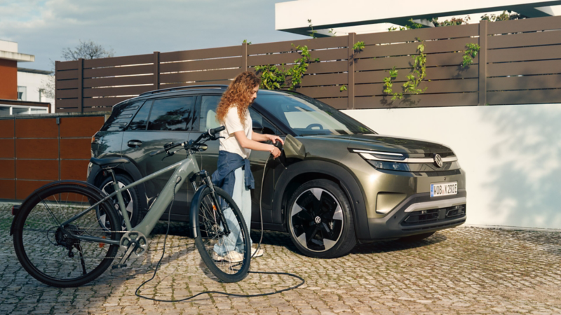 Una mujer está cargando una bicicleta eléctrica mediante un cable desde un VW ID. Cross verde, con una casa residencial al fondo.