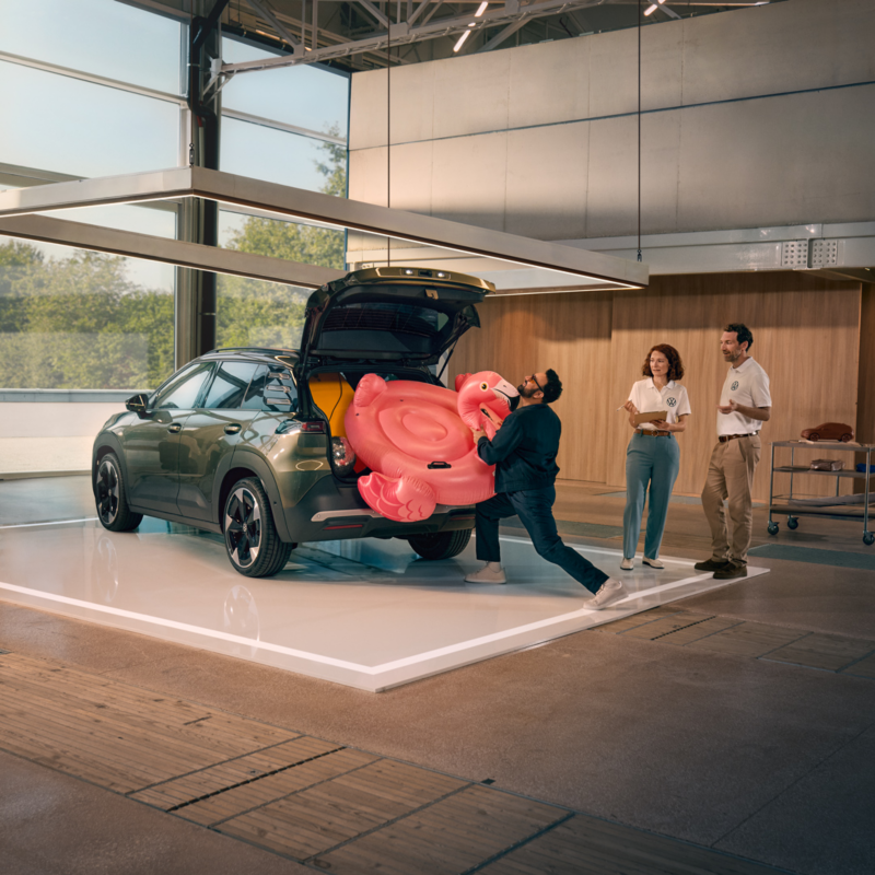 Un homme pousse une bouée gonflable en forme de flamant rose dans le compartiment à bagages ouvert d’un VW ID. Cross. Deux personnes se tiennent à côté du véhicule et observent la scène avec amusement.