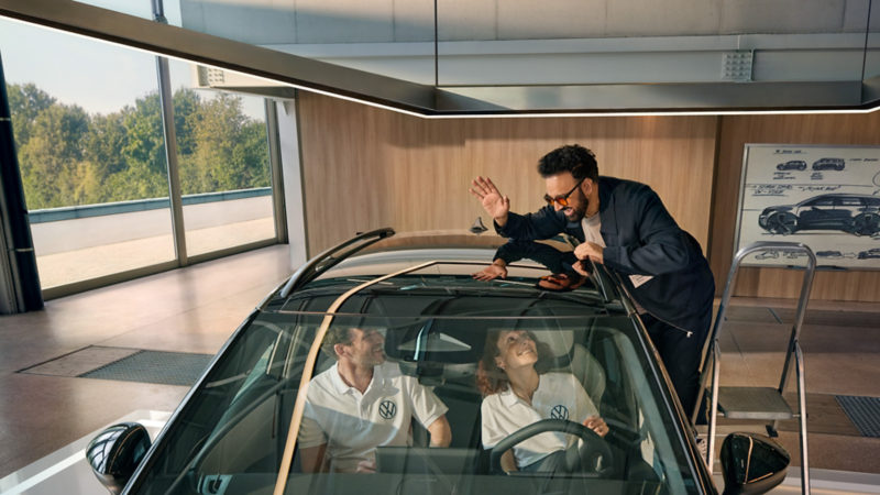 Ein Mann steht auf einer Leiter neben einem VW ID. Cross und winkt durch das Panoramadach den innen sitzenden Personen zu.