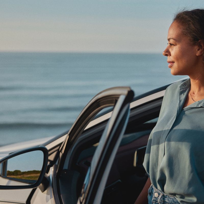 Eine Frau mit langen Haaren steigt aus einem ID.5 von Volkswagen. Im Hintergrund: das Meer.