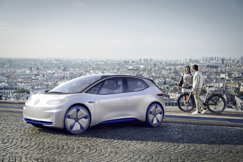 En vit Volkswagen-konceptbil står parkerad på en utsiktsplats med stadsmiljö i bakgrunden, medan två personer med cykel står bredvid.