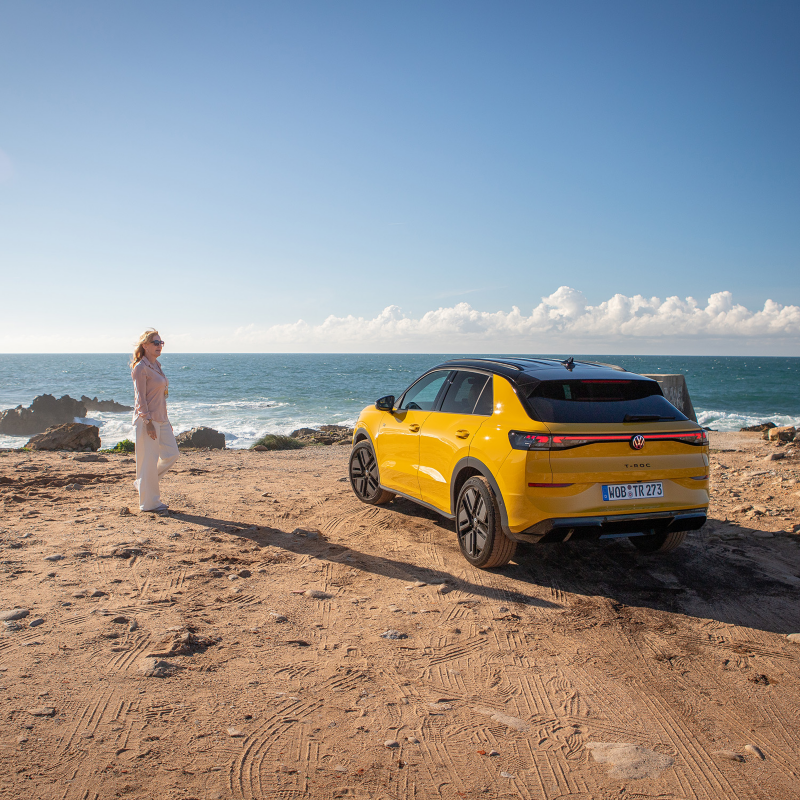 En gul T-Roc är parkerad på en strand vid havet, en kvinna står bredvid bilen