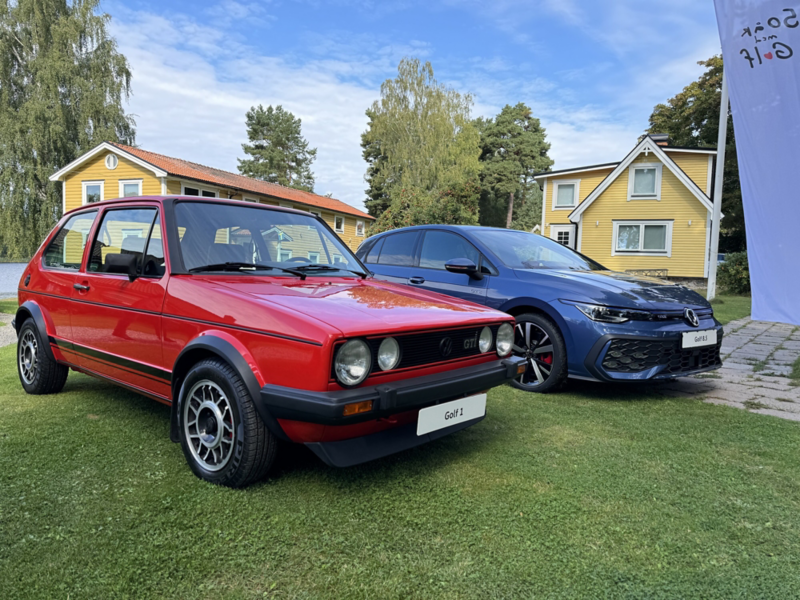 En röd klassisk Volkswagen Golf och en modern blå Volkswagen Golf står parkerade på en gräsmatta framför gula trähus.