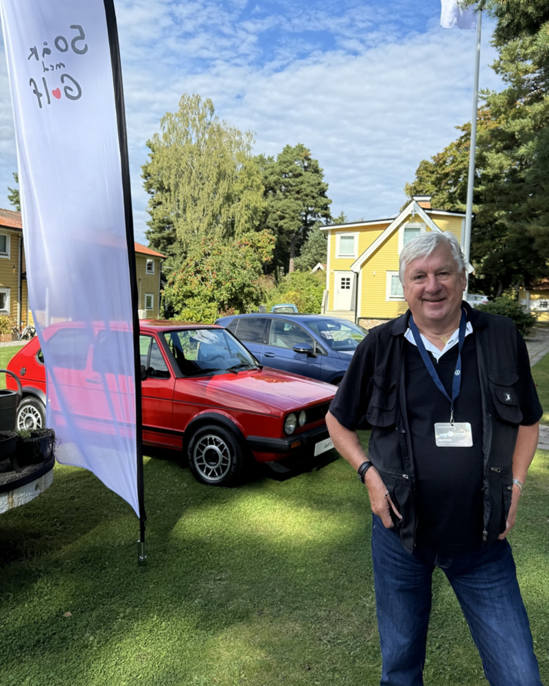 En person står på en gräsmatta, i bakgrunden syns en röd klassisk Volkswagen Golf och en modern blå Volkswagen Golf parkerade