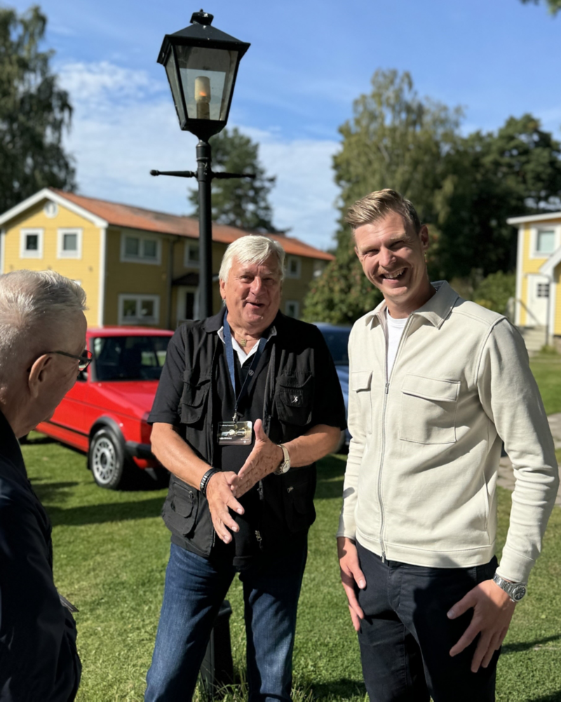 Två personer i framgrunden, i bakgrunden skymtas Volkswagen och gula trähus.
