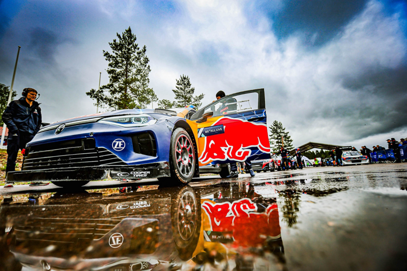 Rallybil parkerad på våt asfalt inför tävling, med team och publik i bakgrunden.