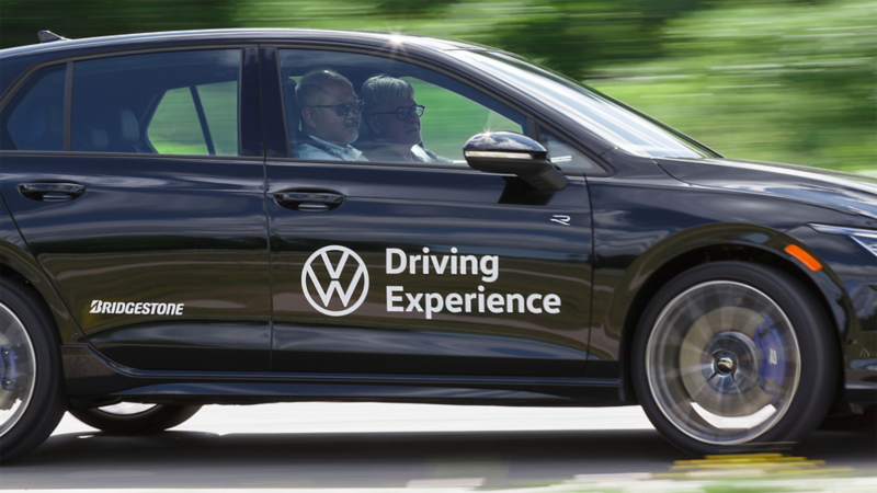 Une voiture Volkswagen noire avec le marquage "Volkswagen Driving Experience" et "Bridgestone" sur son côté est en mouvement, avec deux personnes visibles à l'intérieur. L'arrière-plan est flou pour indiquer la vitesse.