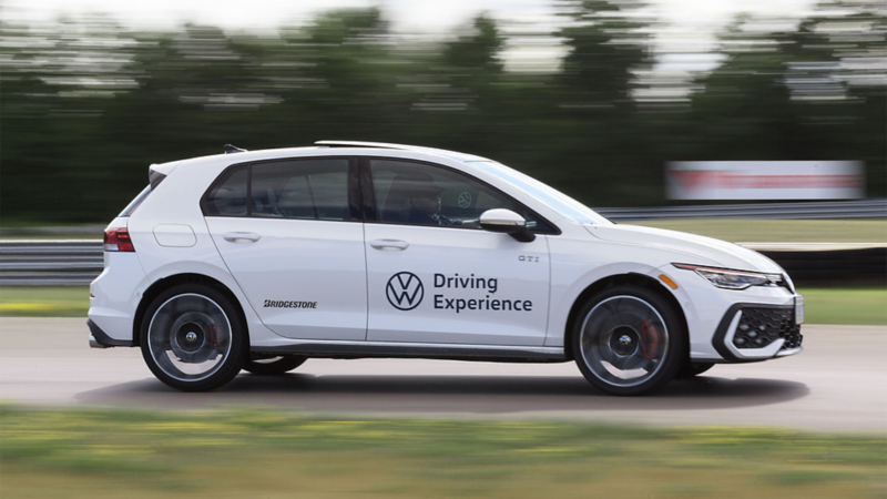 Une voiture Volkswagen GTI blanche avec le marquage "Driving Experience" est en mouvement sur une piste, avec un arrière-plan vert flou indiquant la vitesse.
