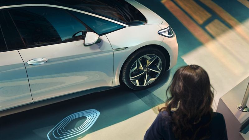 VW ID.3 in Weiss von der Seite sichtbar, Frau geht auf das Fahrzeug zu, Logoprojektion im Fokus