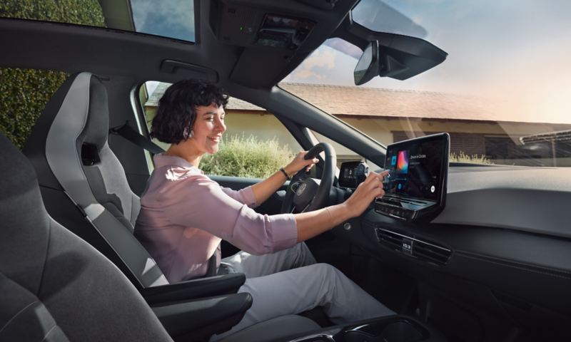 Une femme sourit au volant de sa VW ID.3 en manipulant les commandes de l'écran tactile.
