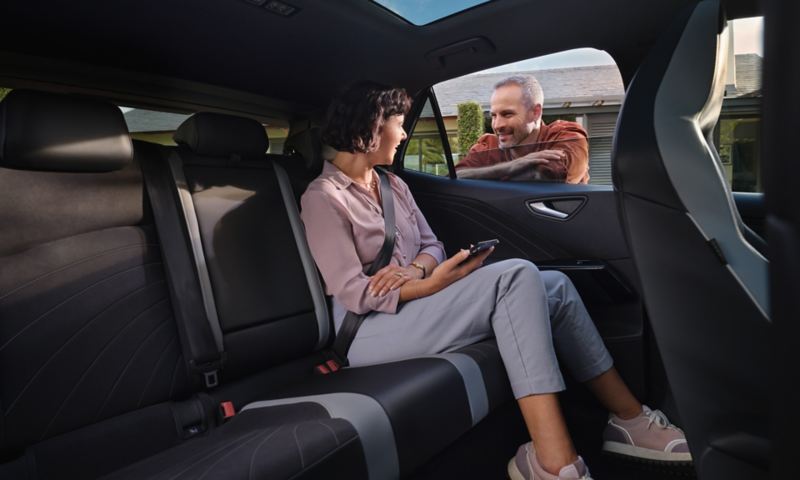 Jeune femme assise, un smartphone à la main, à l'arrière d'une VW ID.3 regarde par la fenêtre un homme lui sourire.