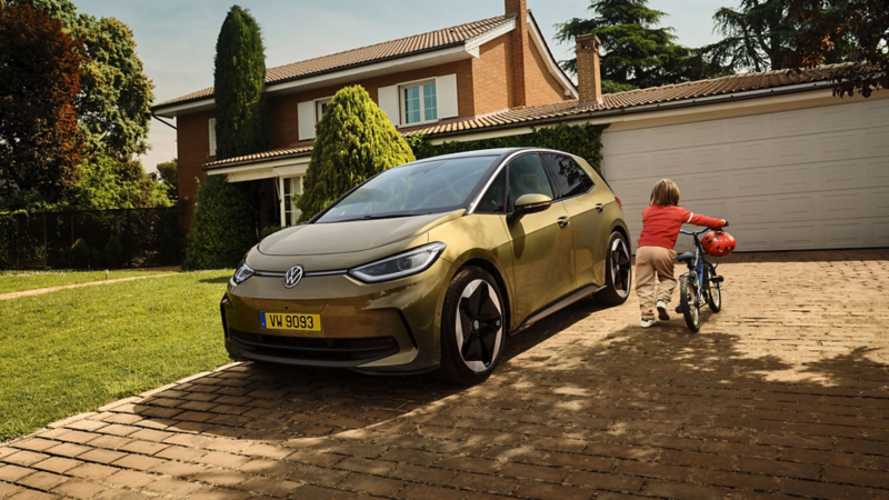 L'ID.3 est garée devant le garage, dans l'allée. À côté, un enfant vêtu d'un pull orange pousse son vélo et son casque vers l'entrée.