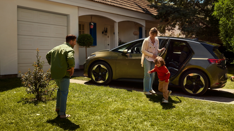 Ein ID.3 steht vor einem Wohnhaus. Die Mutter öffnet die hintere Tür, während ein Kind zum Vater auf dem Rasen läuft.
