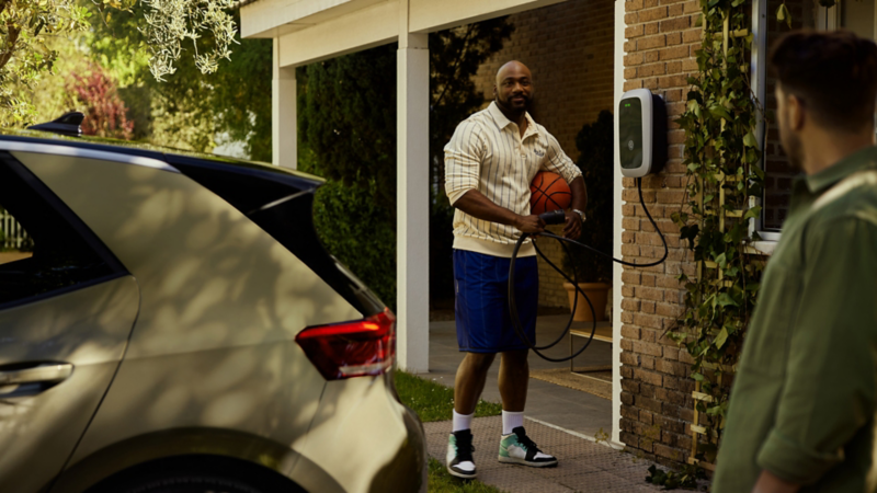  Ein Mann unterhält sich mit seinem Nachbarn im Garten und hält dabei einen Basketball und das ladekabel der Wallbox in der Hand. Im Vordergrund steht ein grüner Volkswagen ID.3