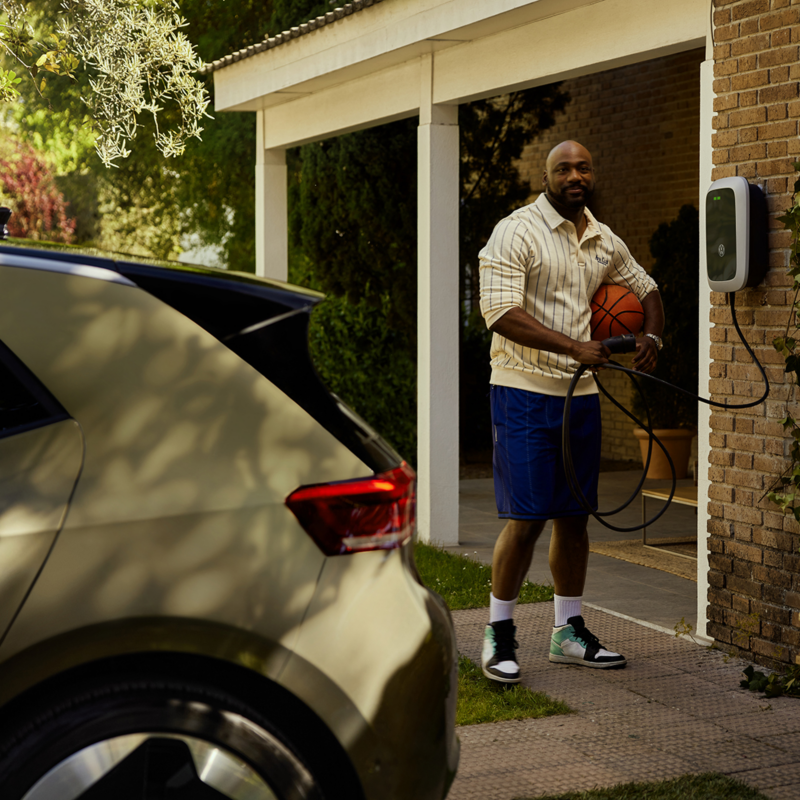 Ein Mann unterhält sich mit seinem Nachbarn im Garten und hält dabei einen Basketball und das Ladekabel der Wallbox in der Hand. Im Vordergrund steht ein grüner Volkswagen ID.3