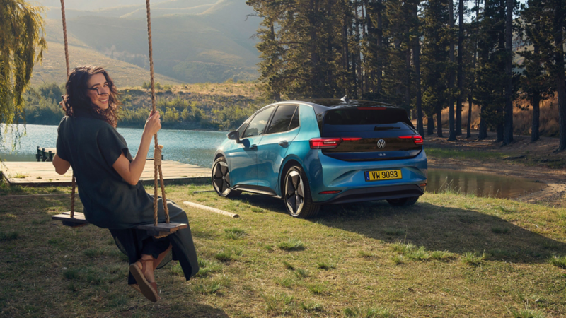Une VW ID.3 bleue est garée au bord d'un lac et une femme est assise sur une balançoire et regarde l'appareil photo. 