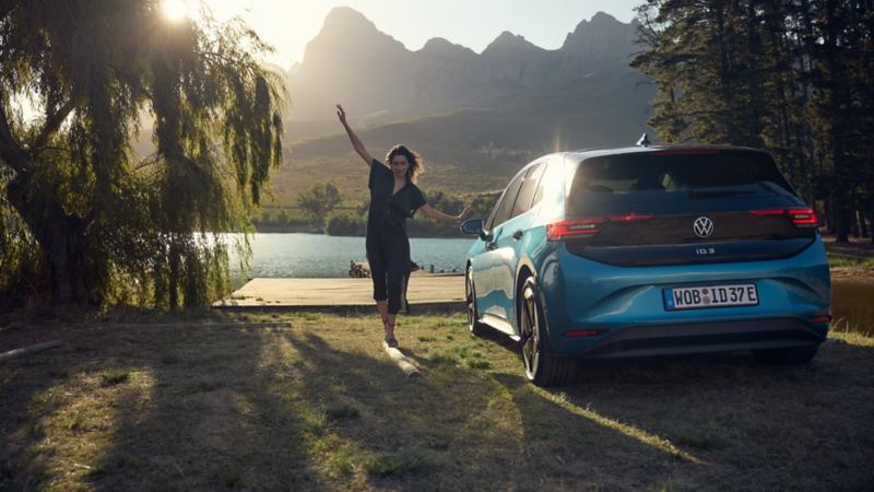 Une femme en équilibre à côté d’une VW ID.3 bleue, vue de l’arrière, devant un paysage de montagne.