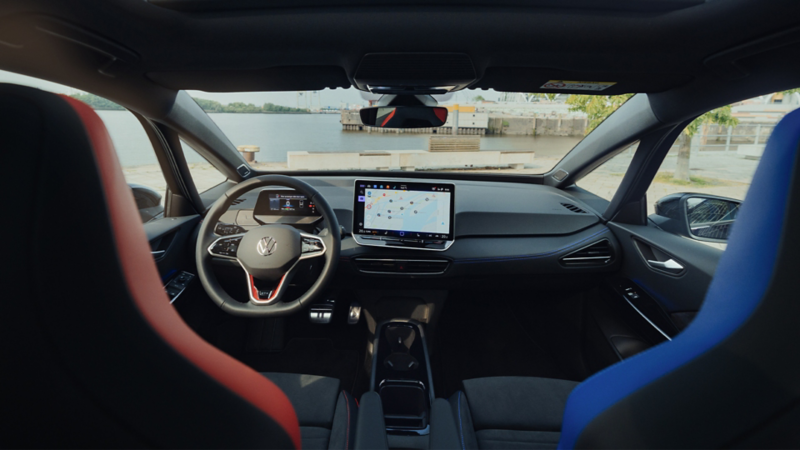  Vue intérieure du tableau de bord d'une Volkswagen ID.3, avec un grand écran de navigation au centre et un petit tableau de bord numérique derrière le volant. Les sièges avant sont en tissu bicolore rouge et bleu. À travers le pare-brise, on voit une vue dégagée sur un port.