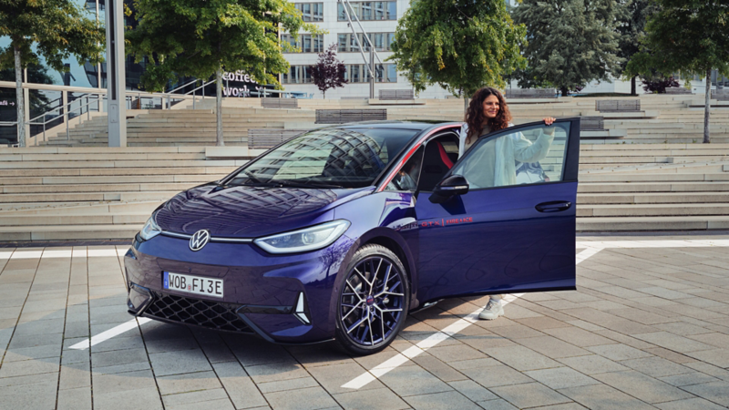 Der ID.3 GTX FIRE+ICE steht auf einer Parkfläche vor einer Betontreppe mit grünen Bäumen im Hintergrund. An der Fahrerseite des ID.s steht eine dunkelhaarige Frau mit sportlicher Jacke.