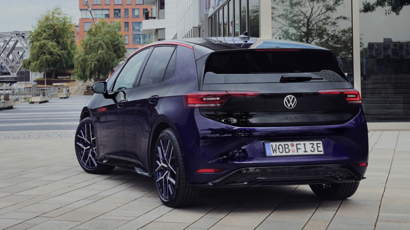 Une VW ID.3 GTX FIRE+ICE violette est garée devant un immeuble de bureaux et un pont ferroviaire.
