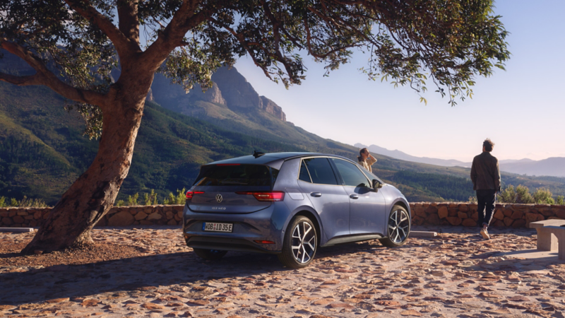 Auf einem Parkplatz in den Bergen parkt ein blauer VW ID.3 Neo, eine Frau und ein Mann genießen die Aussicht.