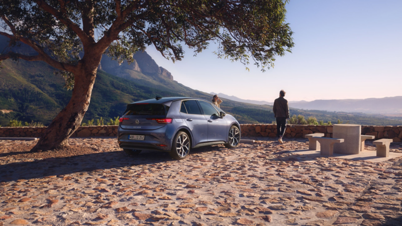 A blue VW hatchback car parked on a cobblestone viewpoint under a tree. Two people stand nearby, gazing at a mountainous landscape under a clear sky.