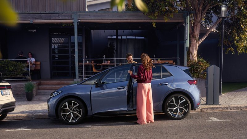 Eine Frau und ein Mann steigen in einen blauen VW ID.3 Neo, der vor einem Café parkt.