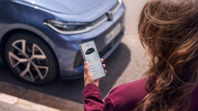 Eine Frau schaut auf ihr Smartphone, vor ihr parkt ein blauer VW ID.3 Neo