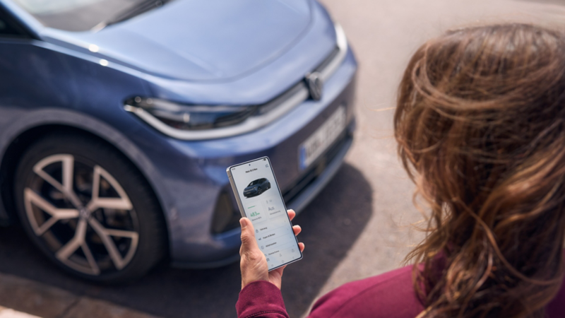 A woman looks at her smartphone with a blue VW ID.3 Neo parked in front of her.