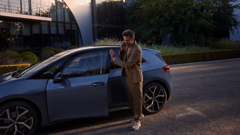 A man stands in front of a blue VW ID.3 Neo and makes a call on his smartphone.