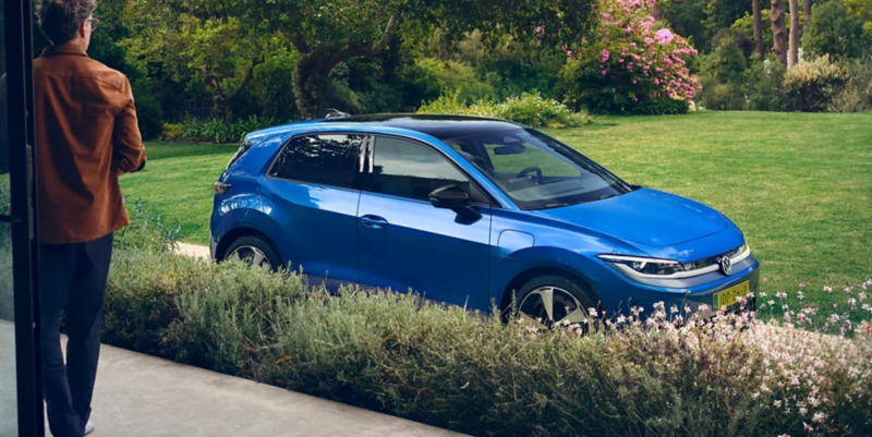 A view through an open terrace door into a green garden, a blue VW ID. Polo stands in the driveway.