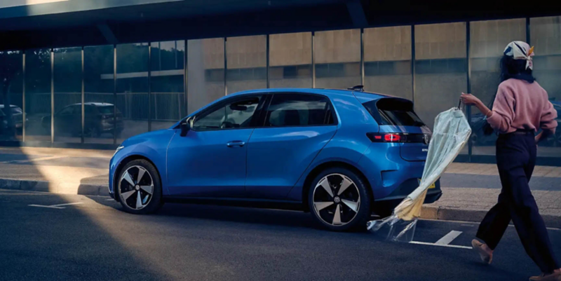 A woman holds her smartphone in her hand, next to her a blue VW ID. Polo is parking driverless backwards into a parking space.