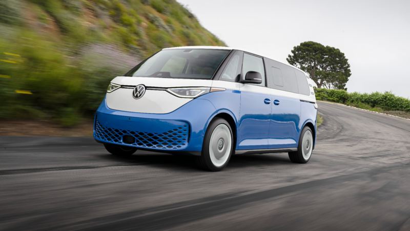 A blue and white 2025 VW ID. Buzz driving on a winding road with green hills and trees in the background.