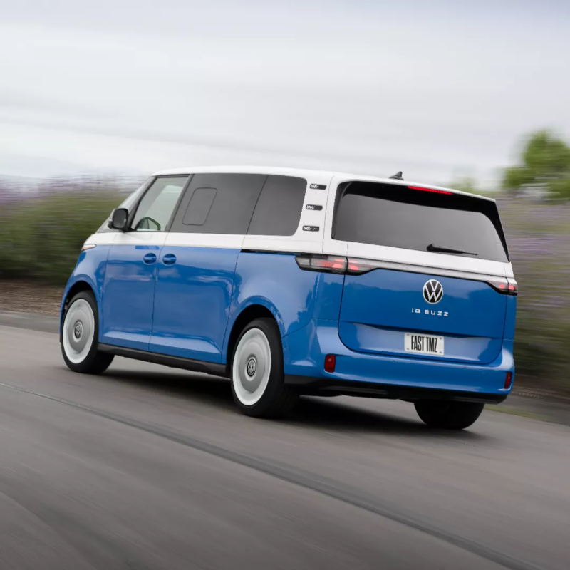 Un Volkswagen ID. Buzz 2025 bleu et blanc roule sur une route sinueuse avec des collines verdoyantes et des arbres en arrière-plan.