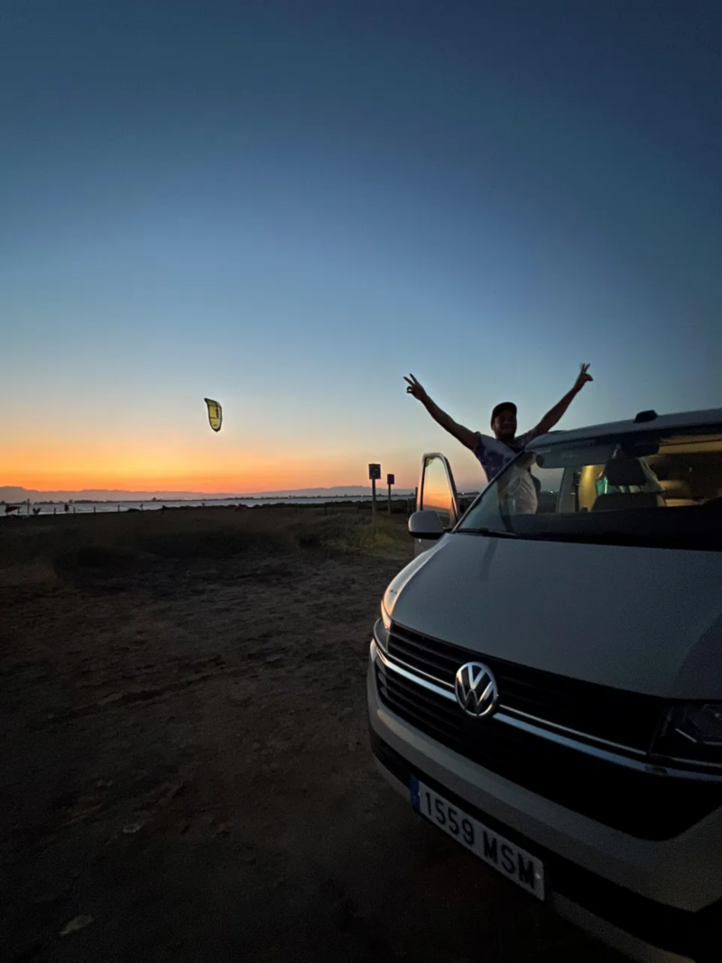 Un VW California en la orilla de una playa con el atardecer de fondo.