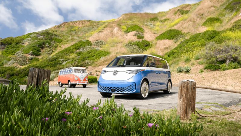 A blue and white Volkswagen ID. Buzz 2025 parked near a hillside with greenery.