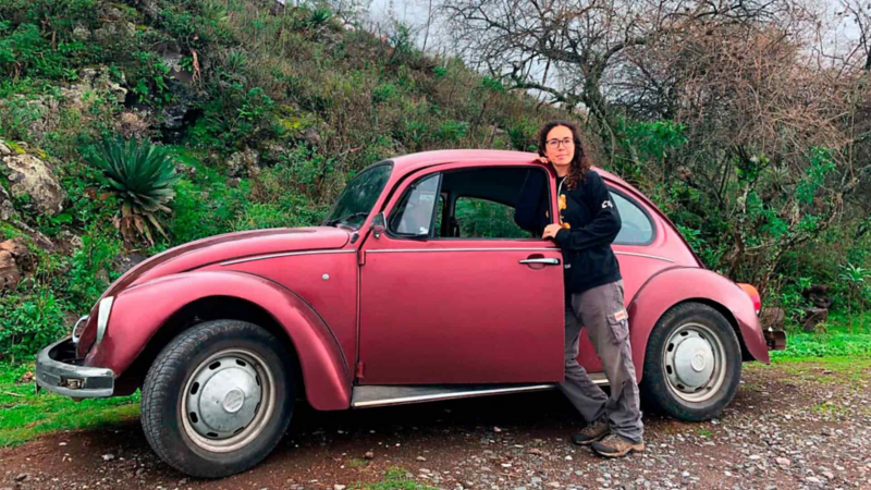 Imagen de Volkswagen Sedán modelo 2000 en color rojo con propietaria en la puerta de piloto.