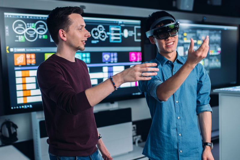 Zwei junge, männliche Kollegen stehen vor großen Displays und experimentieren mit einer VR-Brille