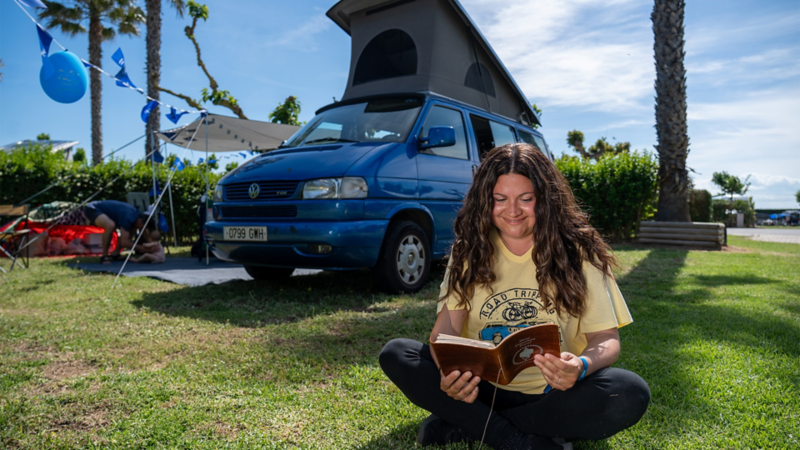 Una mujer sentada en el césped leyendo un libro delante de un VW Multivan.