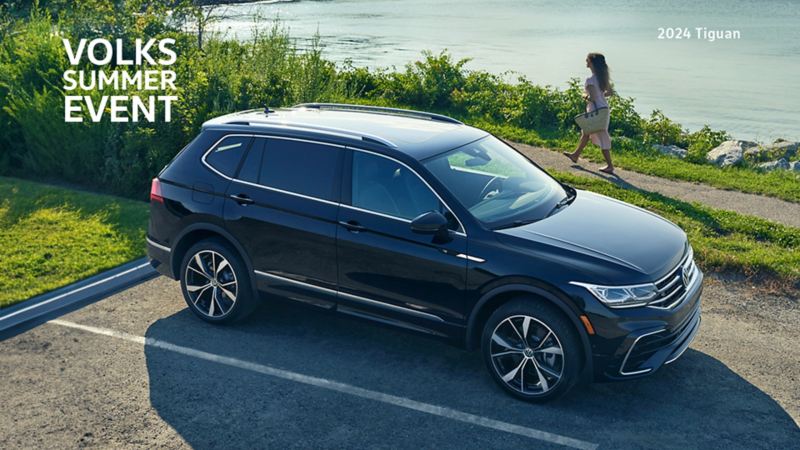 A Tiguan Highline R-Line in Deep Black Pearl is parked on the side of a road, next to a walking path. The path curves around a large body of water, adorned by greenery under a clear, sunny sky. A woman strolls along the path beside the car, enjoying the surroundings.